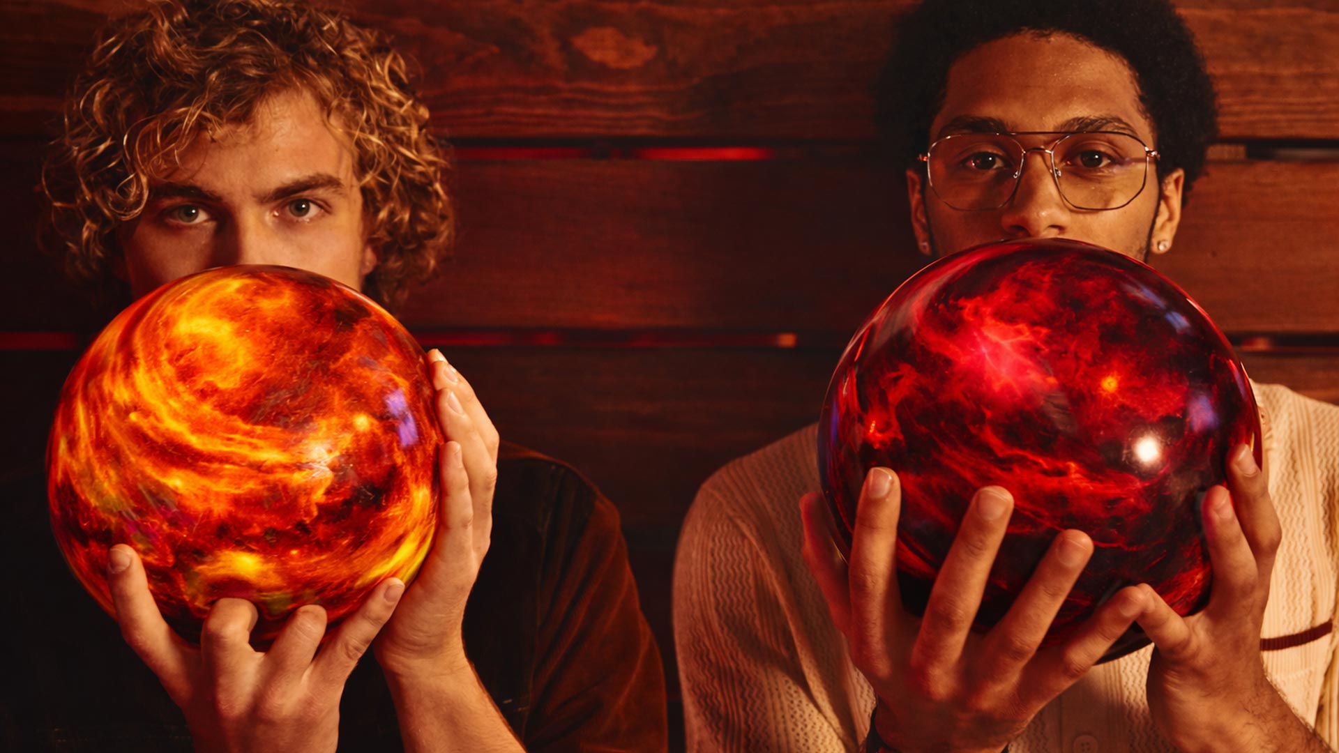 Two young men holding bowling balls in front of their faces, staring directly into the camera with a dramatic, warm-toned background.