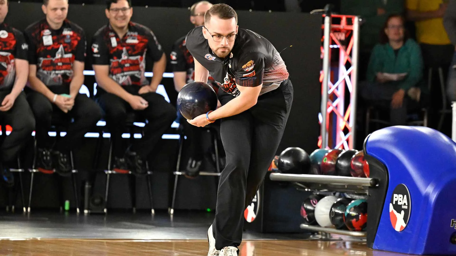 Professional bowler delivering a shot during a PBA competition, with other players seated behind the lanes.