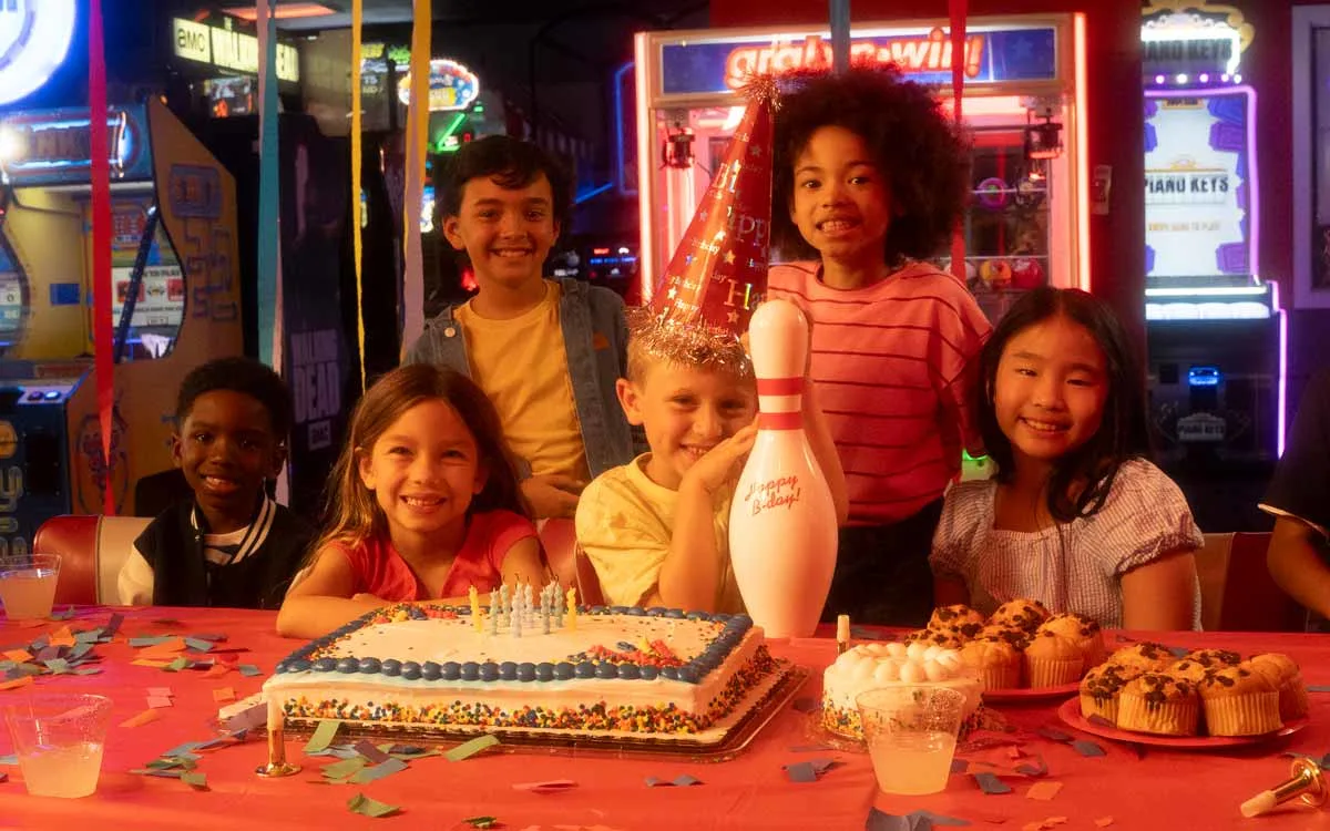 A group of kids sitting behind a table filled with birthday cake, muffins, and a celebratory bowling pin at a kids birthday party at an AMF bowling center