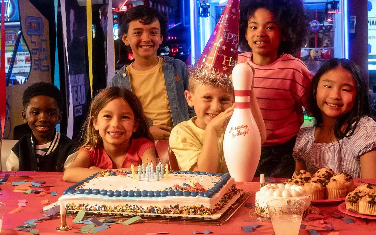 A group of kids sitting behind a table filled with birthday cake, muffins, and a celebratory bowling pin at a kids birthday party at an AMF bowling center