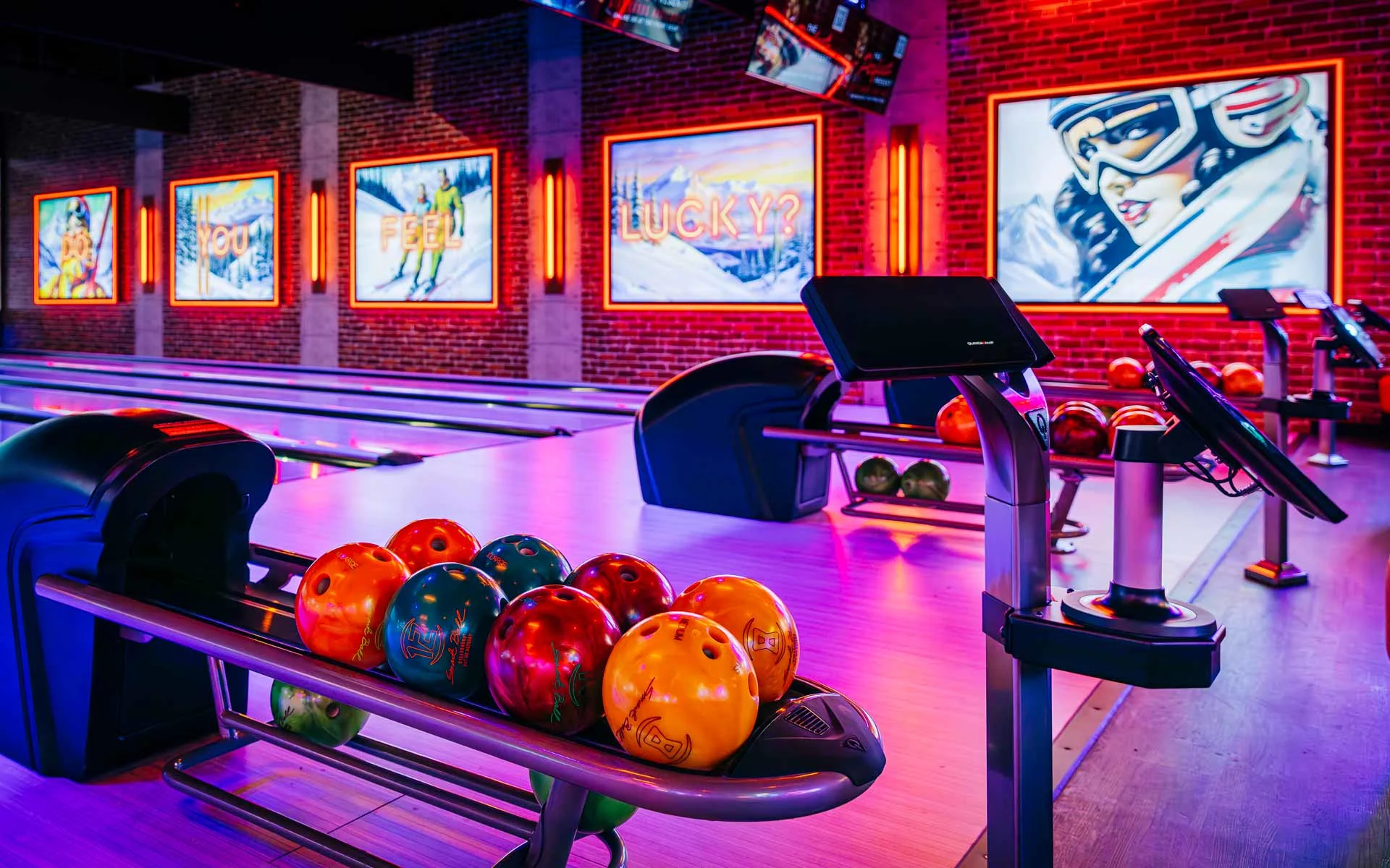 Colorful bowling balls lined up on a ball return beside glowing bowling lanes and scoring screens inside a neon-lit Lucky Strike venue.