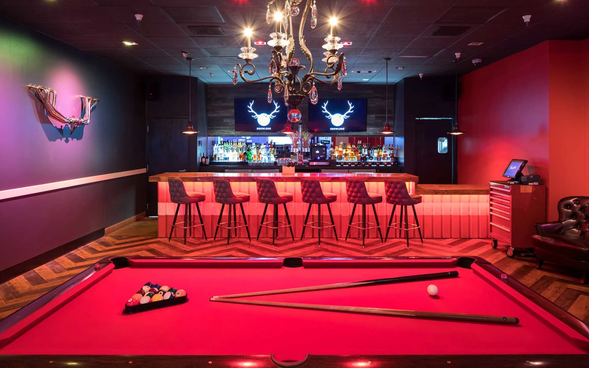 Upscale lounge featuring a red-felt pool table in the foreground, a glowing bar with tufted stools, and a statement chandelier overhead, set against moody lighting and a modern nightlife atmosphere.