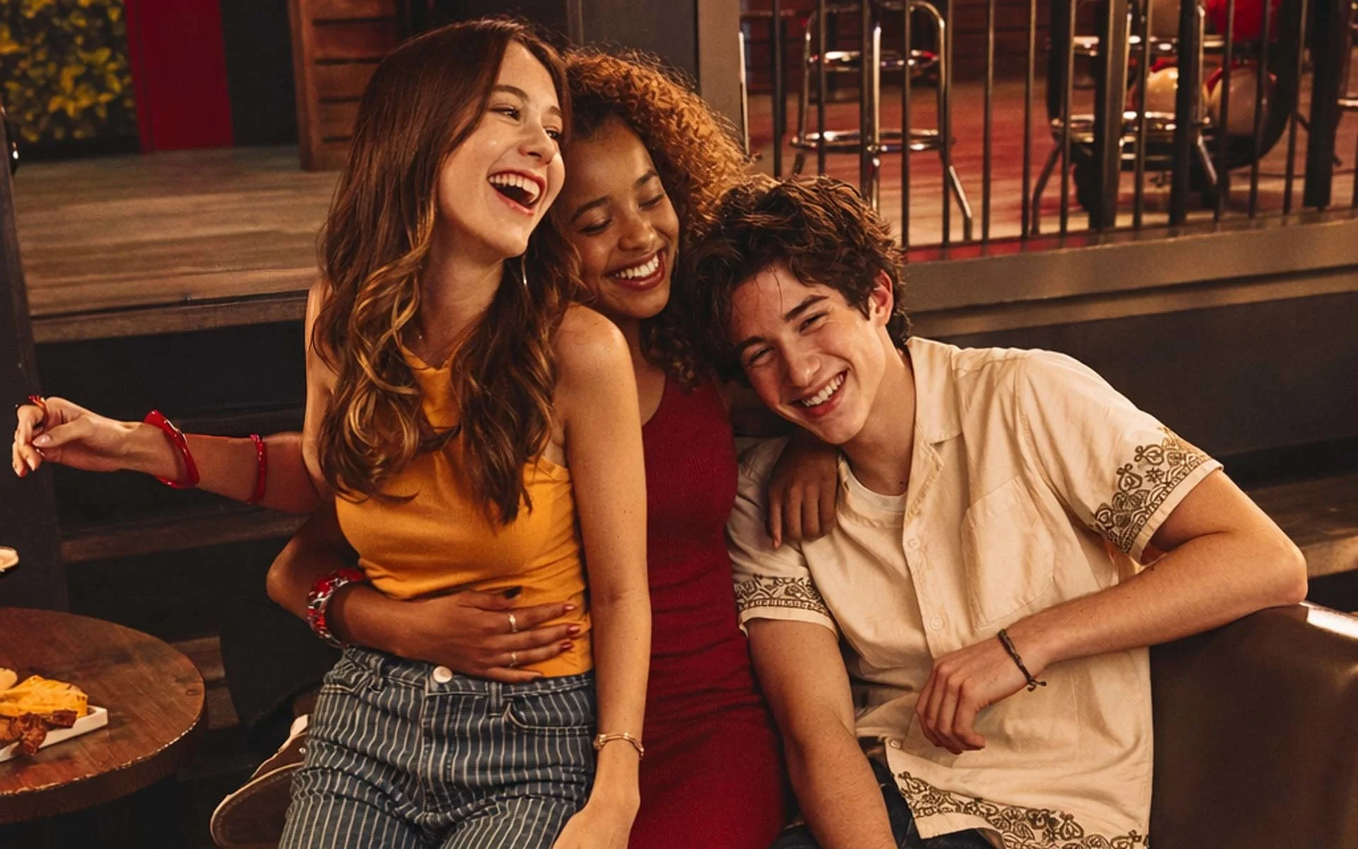 Teenagers laughing together in a modern bowling alley lounge, sitting on a red booth with food and drinks nearby, showcasing a fun, social nightlife atmosphere at an upscale bowling entertainment venue.