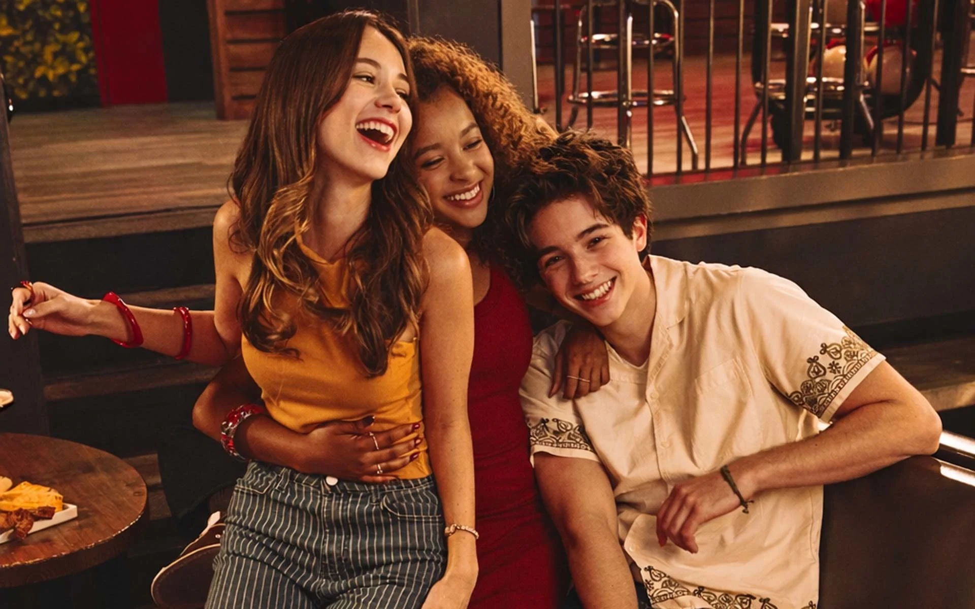 Teenagers laughing together in a modern bowling alley lounge, sitting on a red booth with food and drinks nearby, showcasing a fun, social nightlife atmosphere at an upscale bowling entertainment venue.
