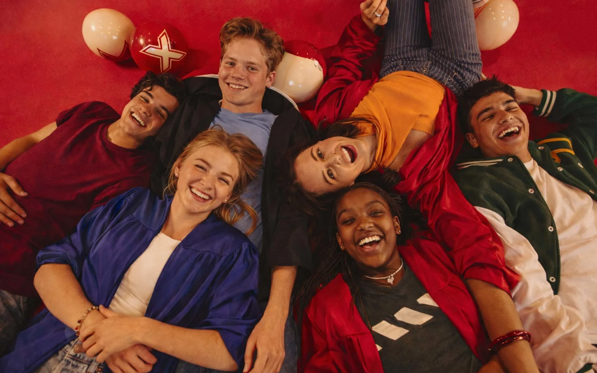 Six teenagers lying in a circle on a bright red floor, smiling and laughing together, with red and white bowling balls around them; one person in the center looks up at the camera while others relax in colorful casual outfits.