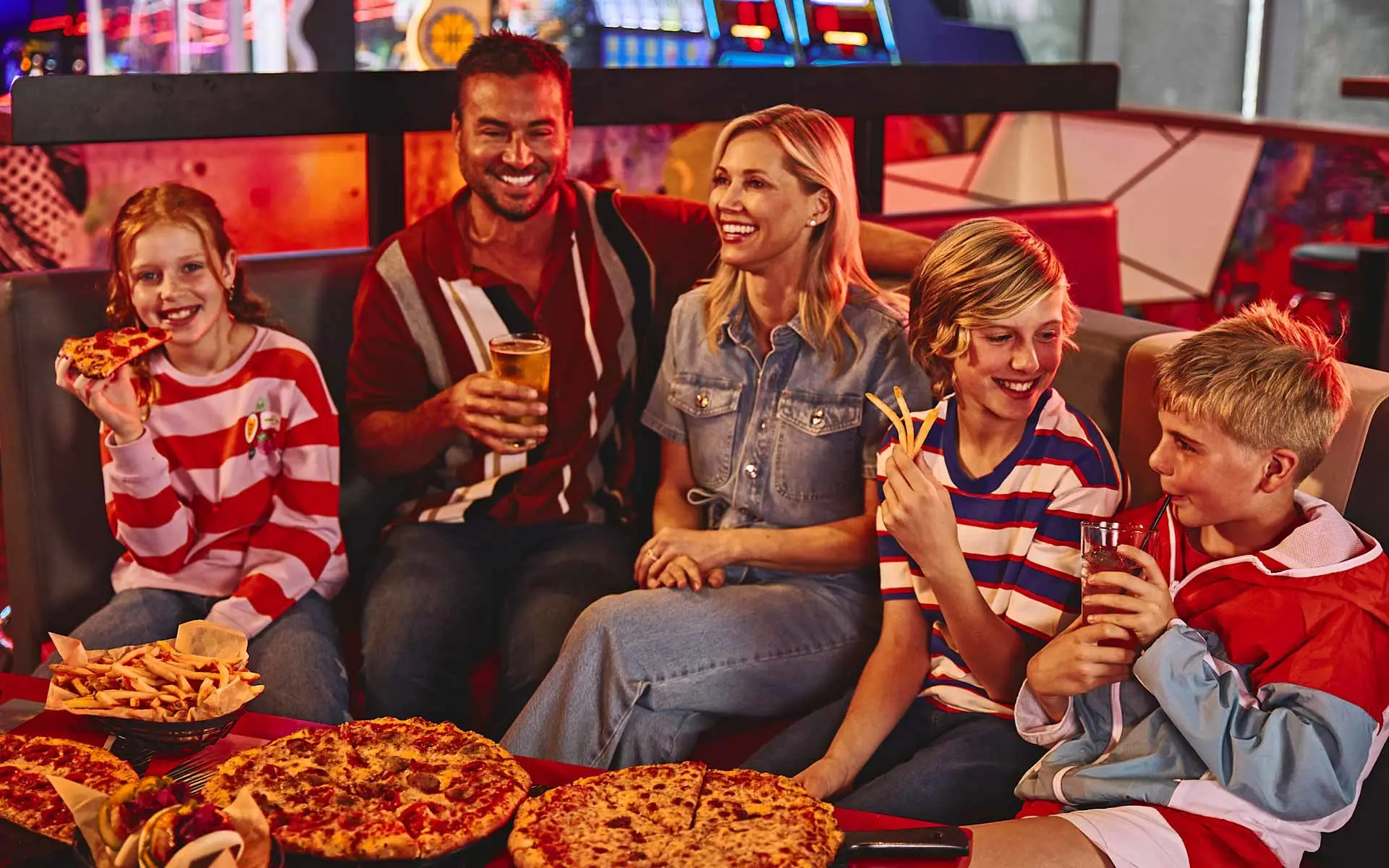 Family smiling and laughing together in a warm, modern bowling lounge with food and drinks on the table.