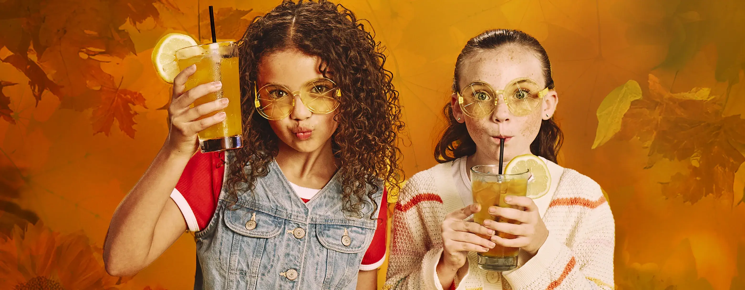 Two girls with caramel apple lemonades in front of an autumn-themed background. 