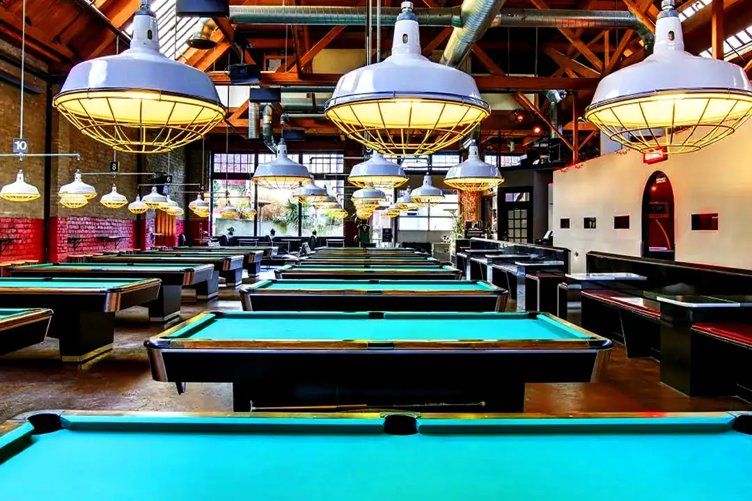 Billiards hall with multiple pool tables arranged under modern white pendant lights and wooden ceiling beams.