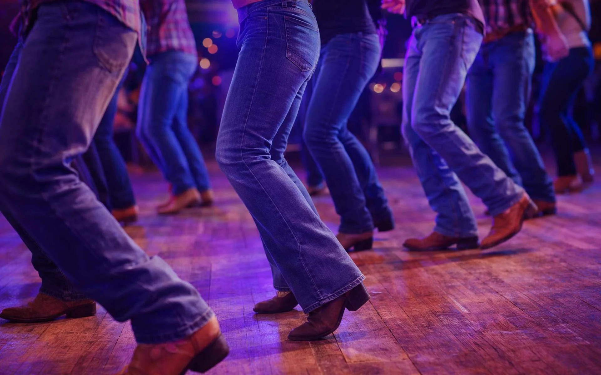 Group of people line dancing in cowboy boots and jeans on a wooden floor with colorful neon lighting in a lively entertainment venue.