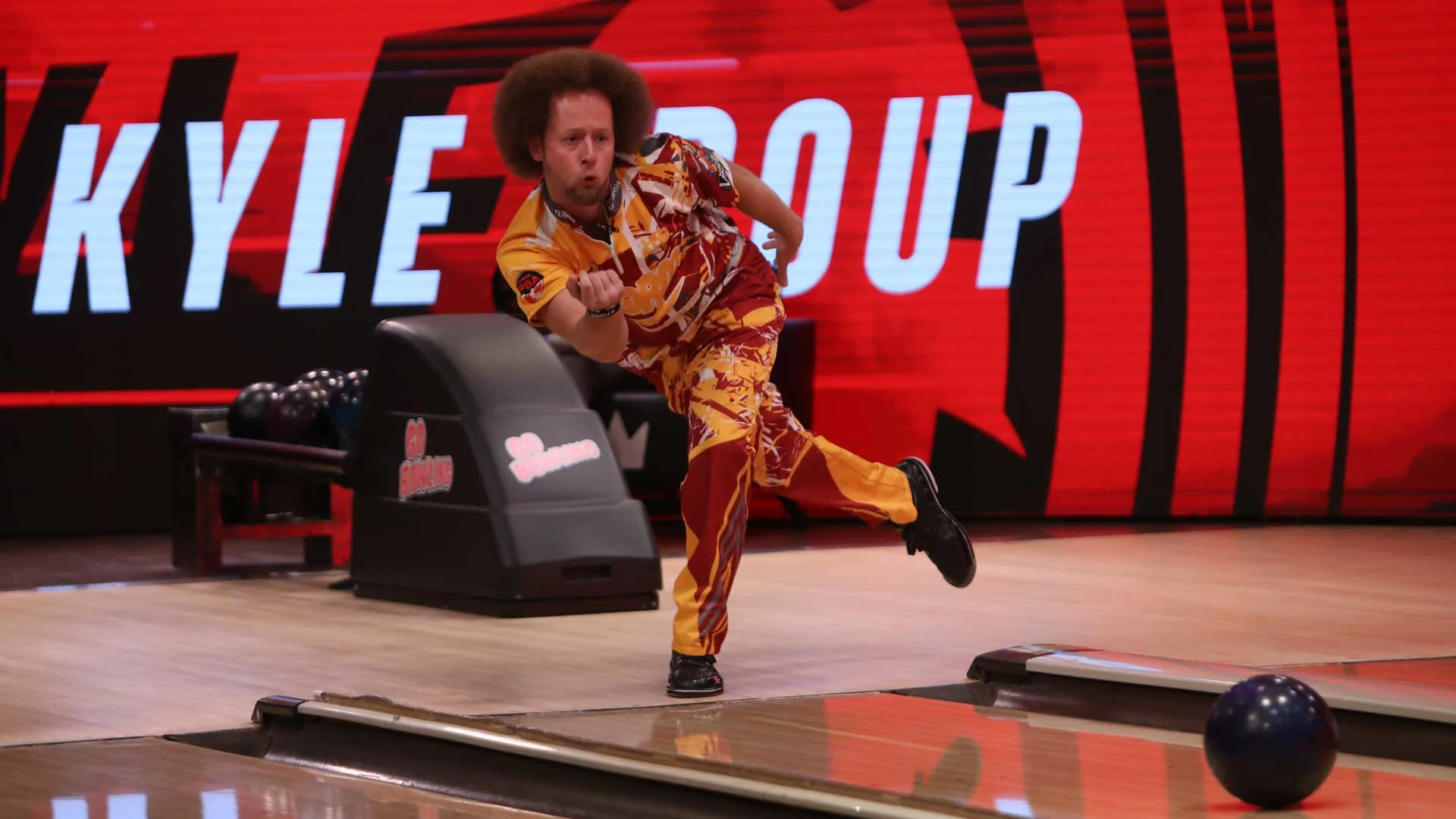 A professional bowler in a bright red and yellow patterned uniform releases a bowling ball down the lane mid-stride, balancing on one leg with focused intensity.