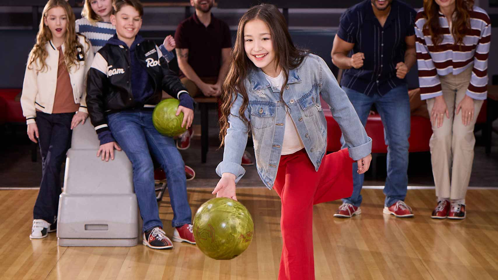 Four kids smiling and holding bowling balls at a bowling center, with adults blurred in the background.