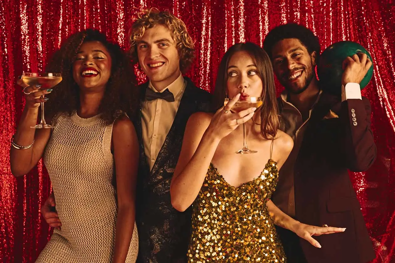 Friends celebrating at a Lucky Strike holiday party, holding cocktails and posing in front of a shimmering red sequin backdrop.