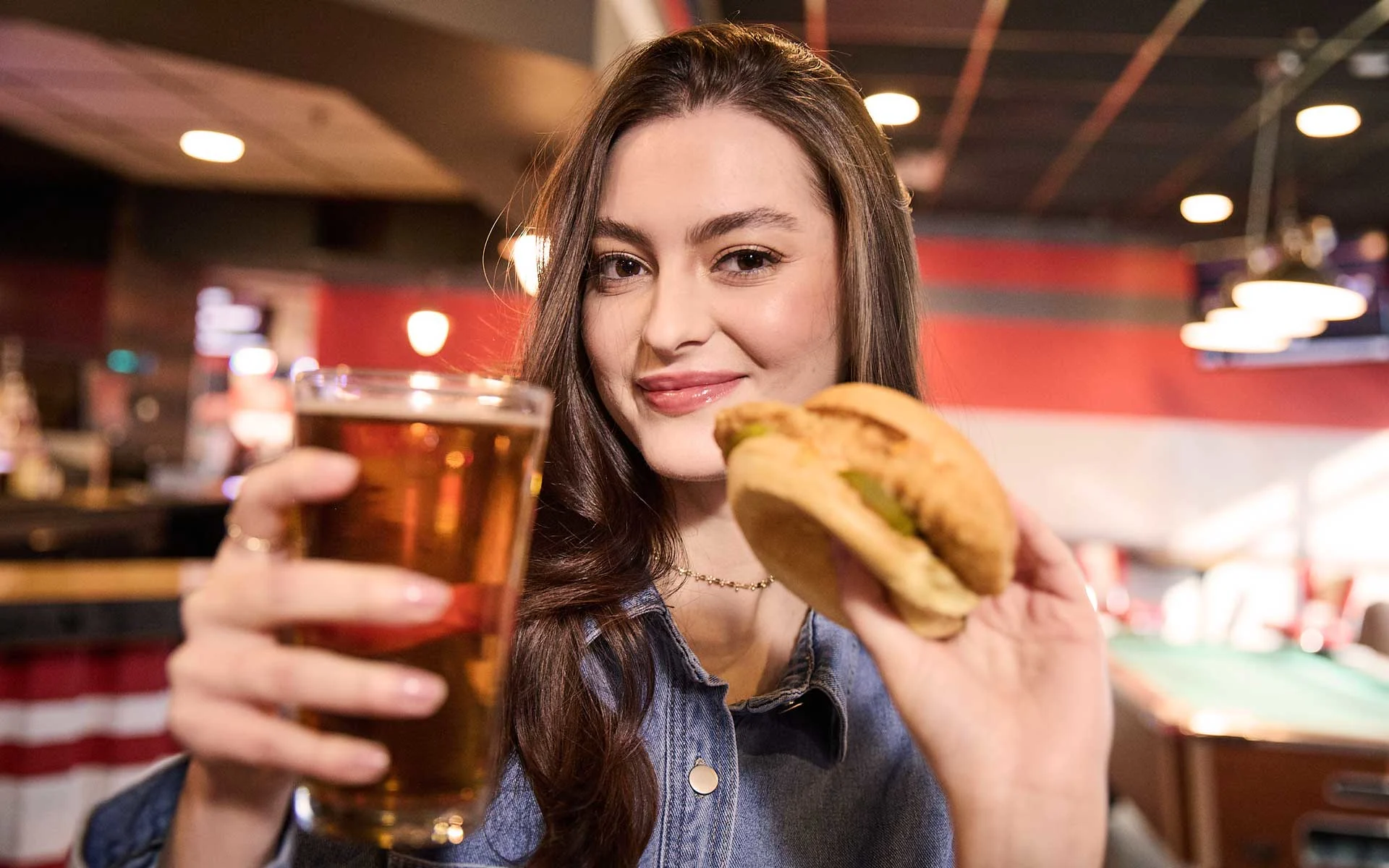 Woman smiling at a bowling alley bar, holding a pint of beer and a crispy chicken sandwich, with warm lighting, pool tables, and a lively entertainment venue atmosphere in the background.