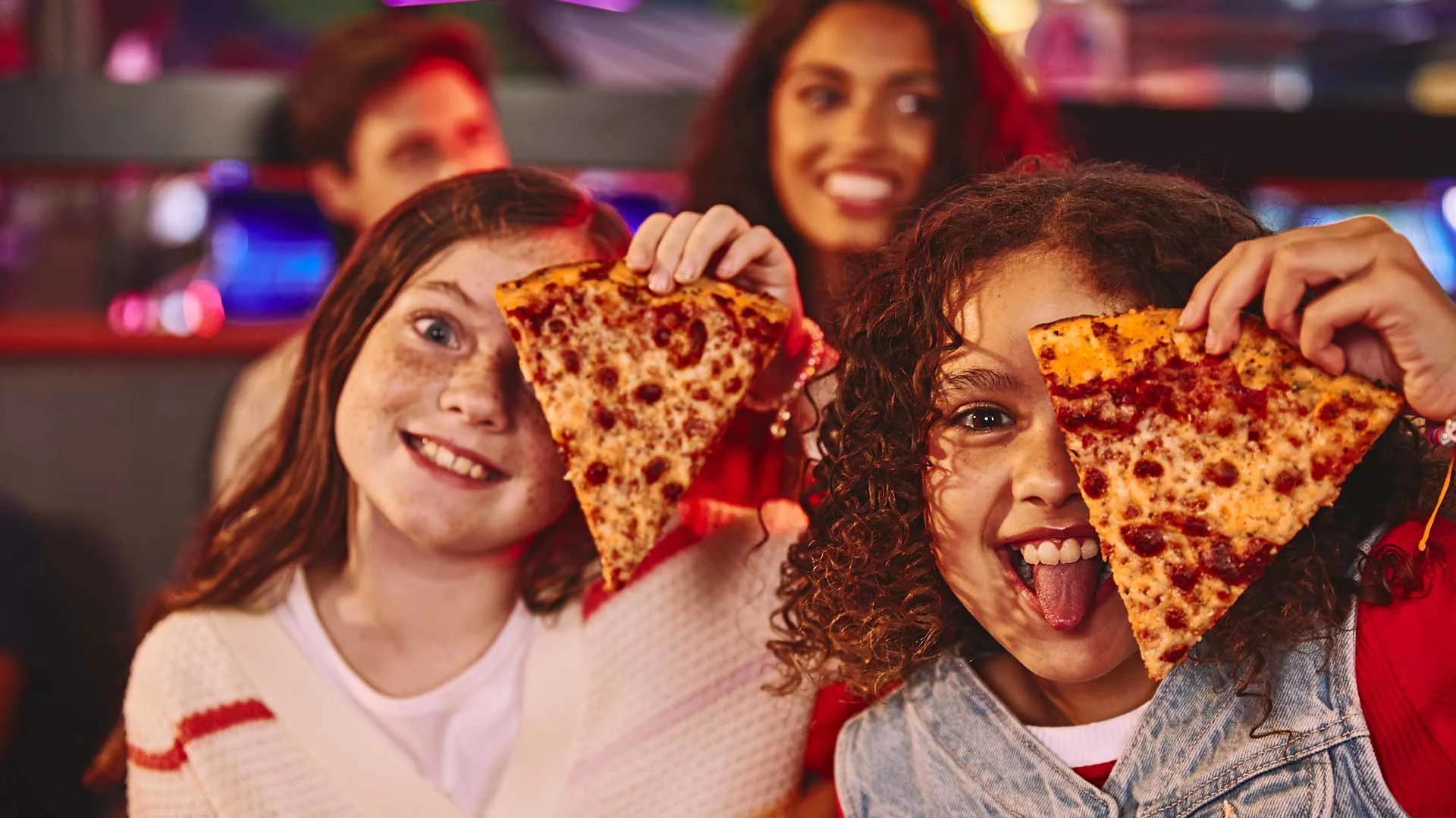 Kids smiling and holding slices of pizza during a fun night out at a bowling center.