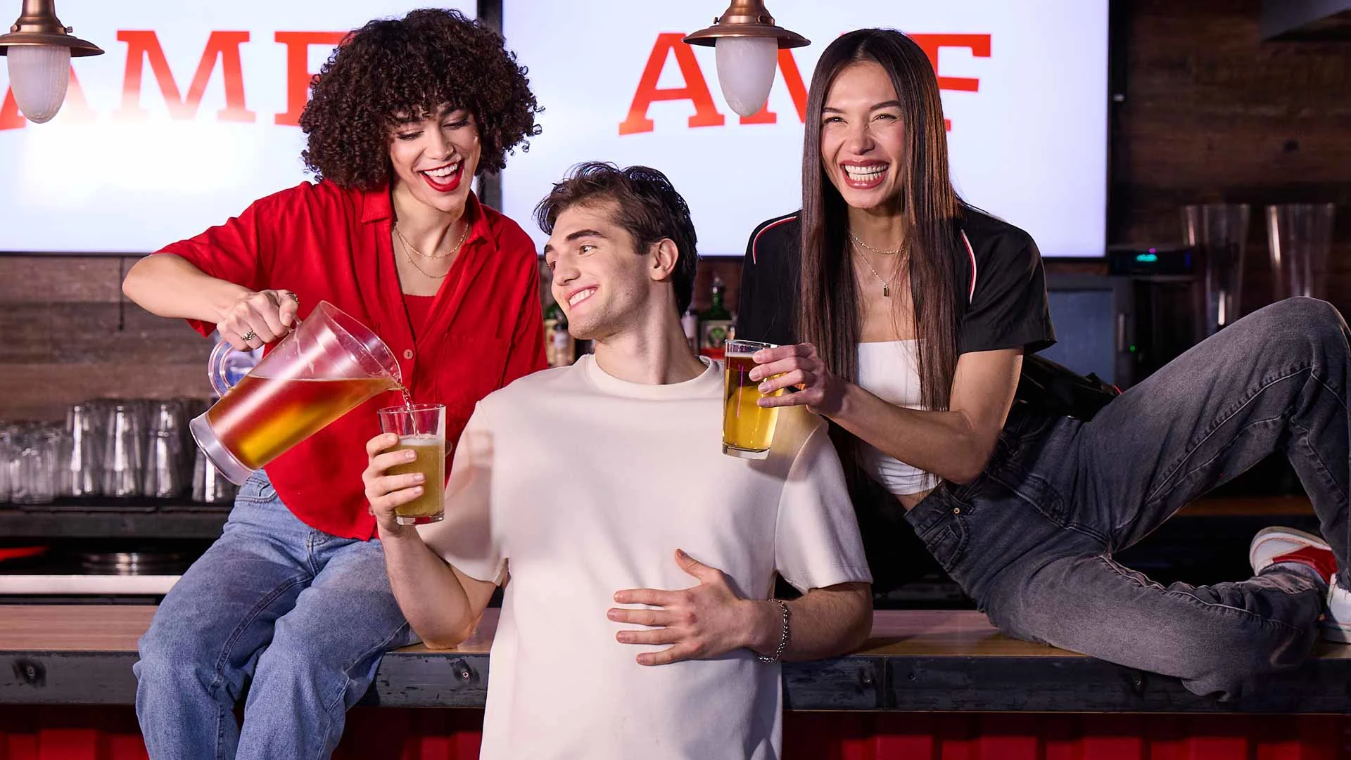 Three friends smiling at a bar, pouring and holding drinks in front of an AMF bowling sign.