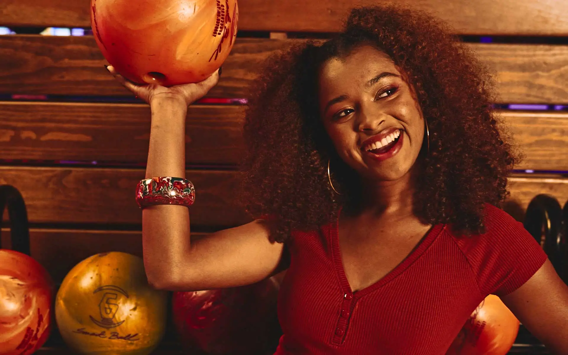 A woman holding a bowling ball above her head while smiling and standing in front of a bowling ball rack at a Lucky Strike bowling alley.