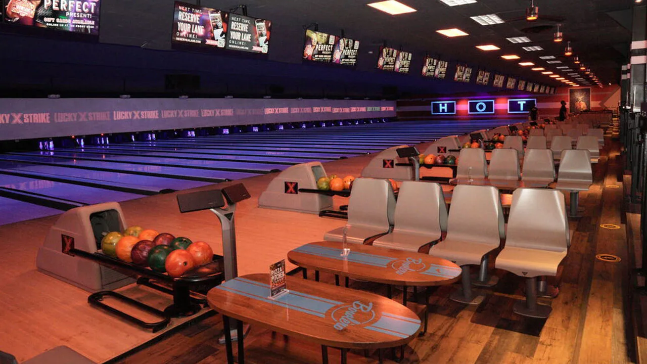 Colorful bowling balls in a return rack with bowling lanes and large TV screens in the background