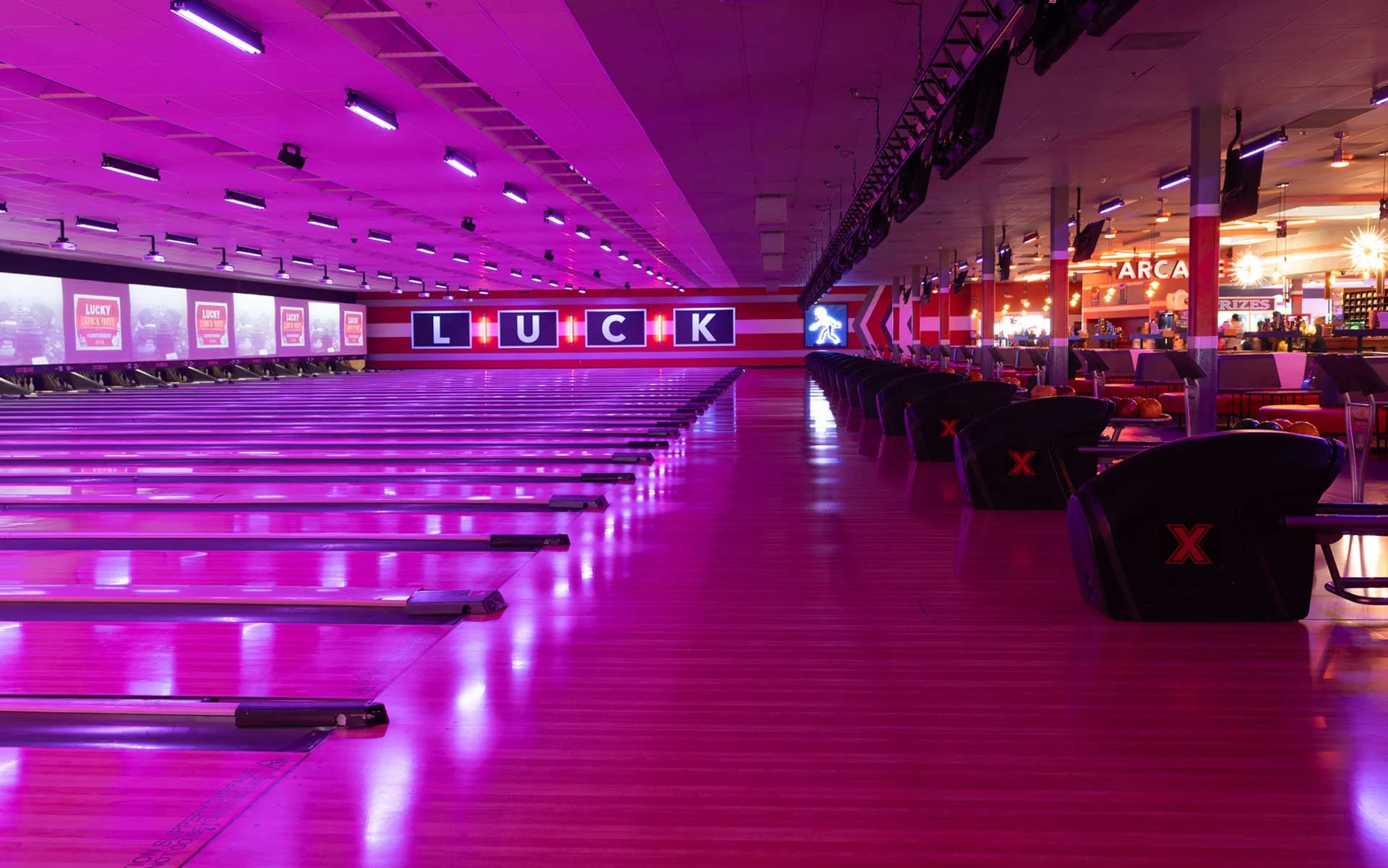 Wide view of a modern bowling center with glowing pink lanes, black seating, overhead scoring screens, and a bright arcade area in the background.