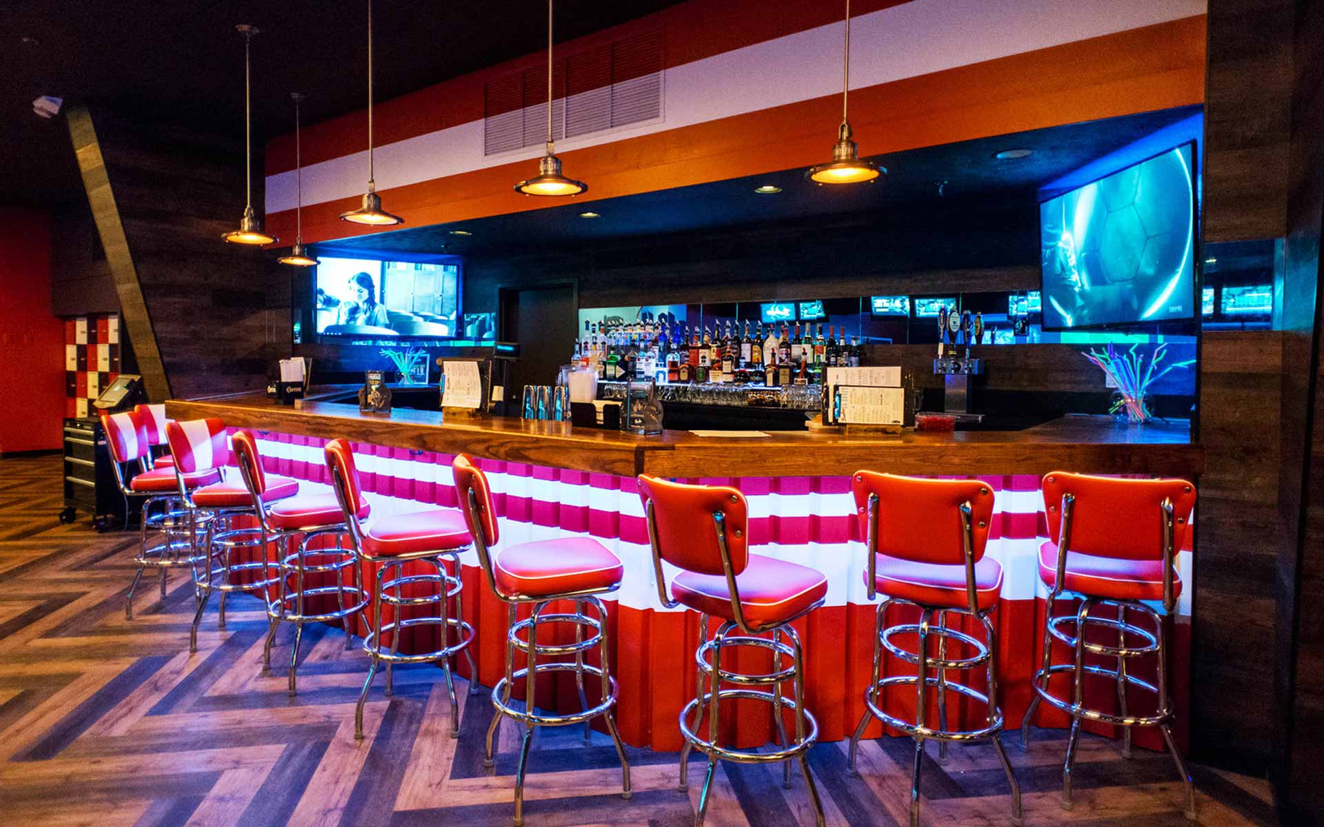 Modern bowling alley bar area with a retro-inspired red-and-white counter, chrome bar stools, and warm pendant lighting, featuring a fully stocked bar, mounted TVs, and a stylish lounge atmosphere.