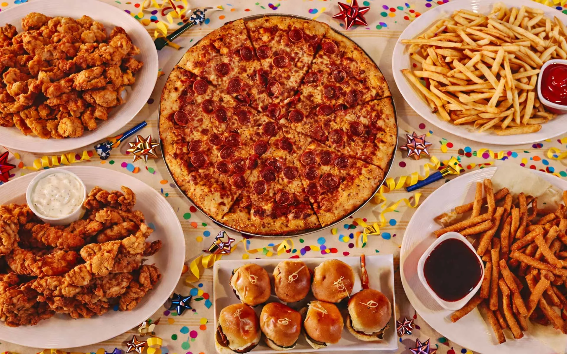 Overhead view of a festive party food spread on a confetti-covered table, featuring a pepperoni pizza at the center surrounded by crispy fried chicken with a side of ranch, golden French fries with ketchup, seasoned fries with dipping sauce, and mini cheeseburger sliders, all arranged on white plates with colorful ribbons and decorations.