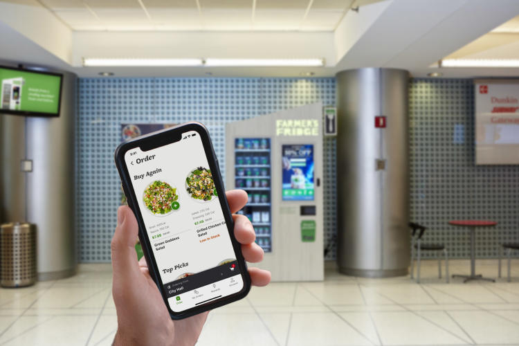 A Farmer's Fridge in a hallway blurred out with a phone in focus showing the app.