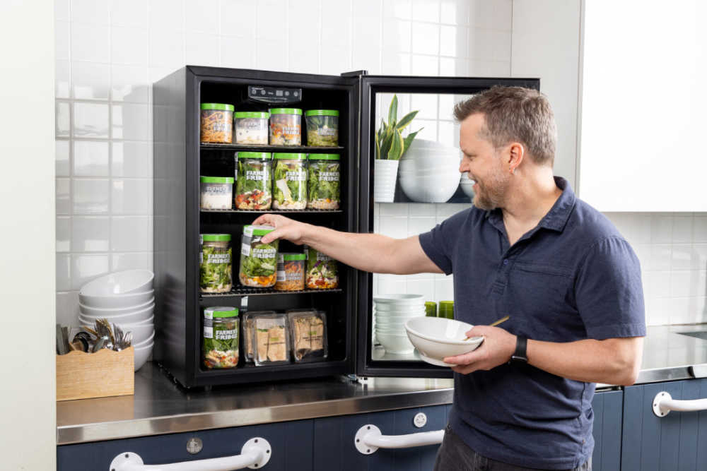 A mini fridge stocked with Farmer's Fridge Jars and a man holding a bowl reaching in to grab one. 