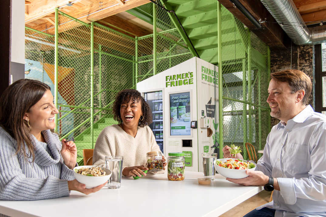 Image of people laughing and eating food, sitting in front of a Farmer's Fridge smart vending machine.