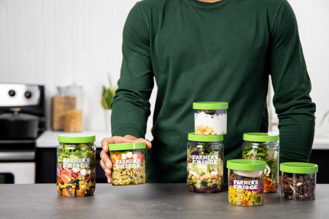 A person holding a Pesto Pasta bowl with other Farmer's Fridge meals on the counter top.