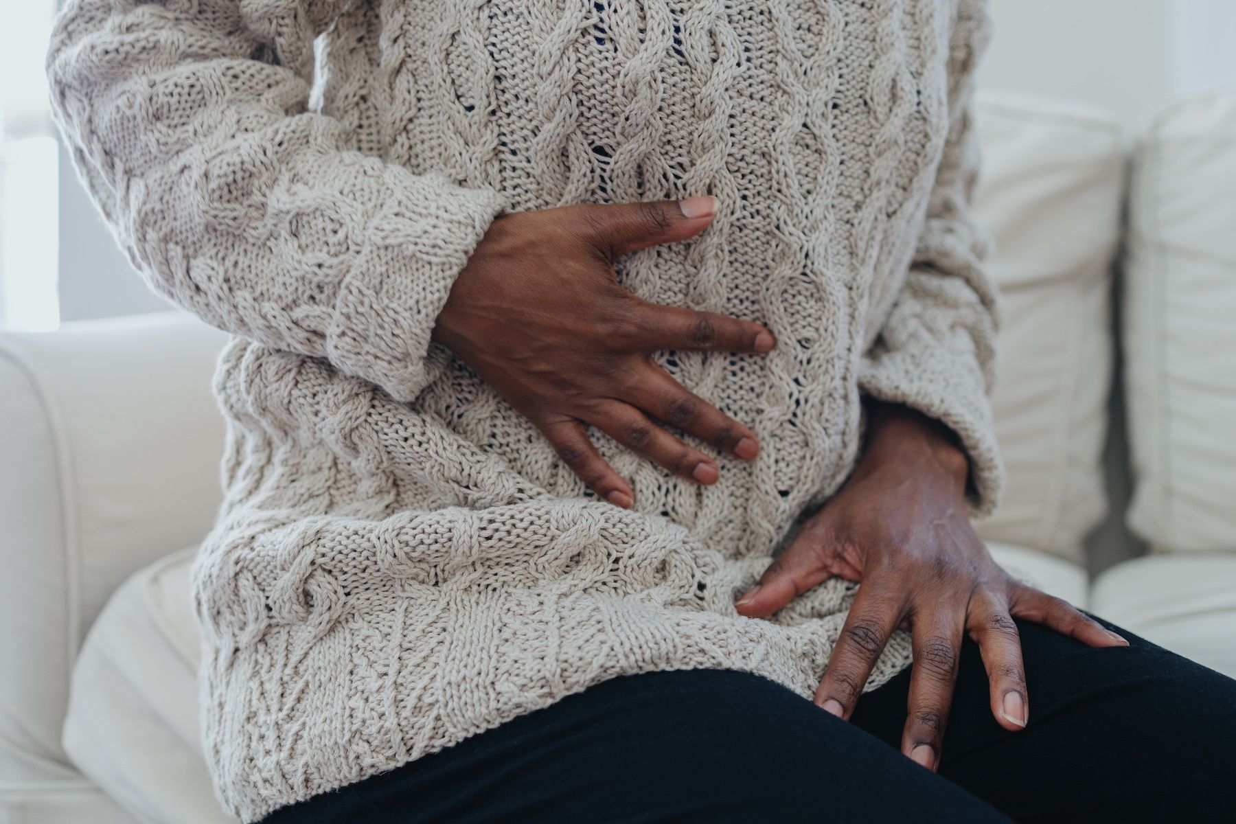The image focuses on a close-up of a person’s torso and hands, conveying a sense of discomfort or pain. The individual, who appears to have dark skin, is seated on a light-colored, possibly white, sofa. They are wearing a chunky, cream-colored cable-knit sweater, which dominates the visual field. The sweater’s texture is prominent, with the intricate cable patterns clearly visible. The person’s hands are clasped over their lower abdomen, with fingers pressing gently but firmly into the area. This gesture strongly suggests abdominal pain or discomfort. They are also wearing dark-colored pants, providing a contrast to the lighter tones of the sweater and sofa. The background is softly blurred, keeping the focus entirely on the person and their physical state. The lighting is soft and natural, creating a somewhat muted and empathetic atmosphere. The composition is intimate, drawing the viewer’s attention to the person’s vulnerability and the physical sensation they are experiencing. The overall impression is one of illness, distress, or a need for comfort. The image evokes a feeling of empathy and concern for the individual’s well-being.