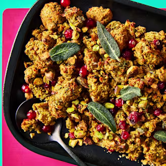 Vegan stuffing in a black serving dish topped with cranberries and sage leaves on a pink and teal background.