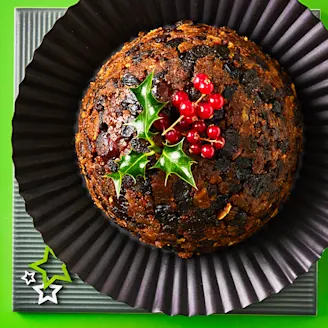 Tofu vegan Christmas pudding topped with holly and berries on a black plate and green background with stars.