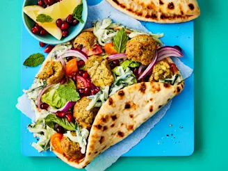 Open bread wraps with crumbled falafel, salad, red onion and chopped tomatoes on a blue board with lemon wedges and pomegranate seeds.