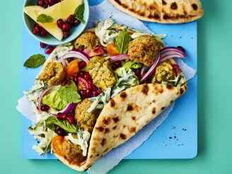 Open bread wraps with crumbled falafel, salad, red onion and chopped tomatoes on a blue board with lemon wedges and pomegranate seeds.