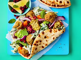 Open bread wraps with crumbled falafel, salad, red onion and chopped tomatoes on a blue board with lemon wedges and pomegranate seeds.