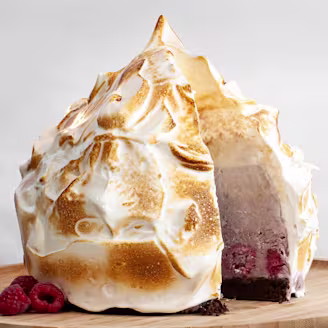 A vegan baked Alaska with a slice removed to showcase the filling with raspberries on the side