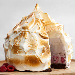 A vegan baked Alaska with a slice removed to showcase the filling with raspberries on the side