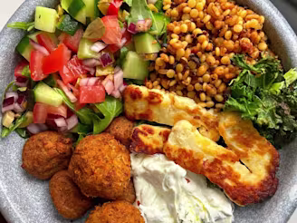A Cauldron Moroccan Falafel Bowl with halloumi, grains, cucumber, tomatoes, onion and mixed leaves with tzatziki on a grey bowl.