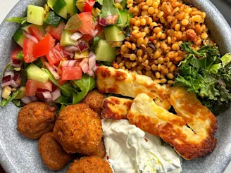 A Cauldron Moroccan Falafel Bowl with halloumi, grains, cucumber, tomatoes, onion and mixed leaves with tzatziki on a grey bowl.