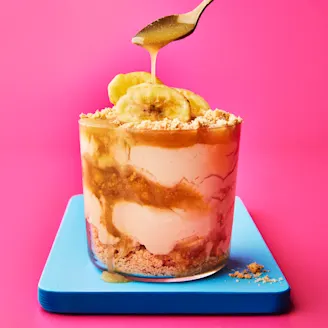 Glass pot with layered sundae filling of biscuit, cream, toffee sauce and banana on blue board with pink background. Spoon drizzling toffee sauce on top.