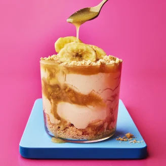 Glass pot with layered sundae filling of biscuit, cream, toffee sauce and banana on blue board with pink background. Spoon drizzling toffee sauce on top.