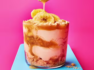 Glass pot with layered sundae filling of biscuit, cream, toffee sauce and banana on blue board with pink background. Spoon drizzling toffee sauce on top.
