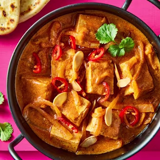 A Butter Tofu Curry with red chilli, almonds and naan bread in a black bowl on a pink background.