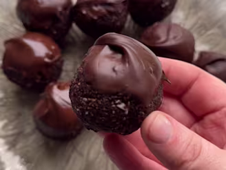 Hand holding a no bake brownie ball made using tofu and dipped in chocolate. Seven more can be seen on a plate in the background.