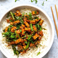Vegan sticky sesame Tofu served with spring greens on a bed of white rice served in a white bowl with chopsticks on the side