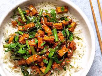 Vegan sticky sesame Tofu served with spring greens on a bed of white rice served in a white bowl with chopsticks on the side
