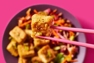 A bowl of Cauldron Extra Firm Tofu bowl with slaw in the background, forefront is a single tofu piece being held my pink chopsticks 