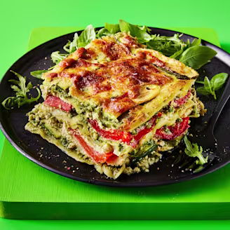 A slice of lasagne with the layers visible to showcase the fillings with a side of rocket served on a dark plate with a green background 
