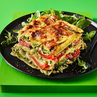 A slice of lasagne with the layers visible to showcase the fillings with a side of rocket served on a dark plate with a green background 