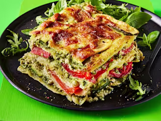 A slice of lasagne with the layers visible to showcase the fillings with a side of rocket served on a dark plate with a green background