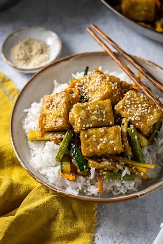 Sticky Teriyaki Tofu served on a bed of rice and vegetables in a white bowl and chopsticks on the side